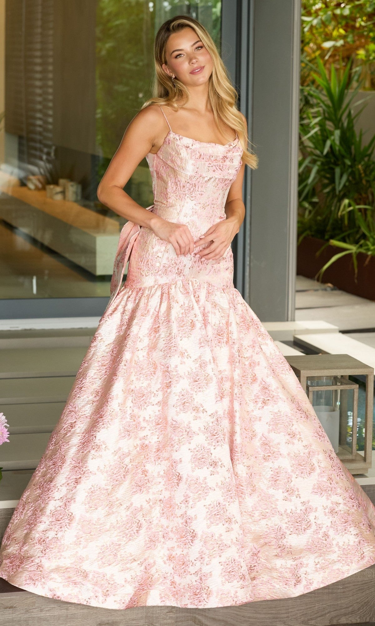 A woman stands outside in front of glass doors, wearing the Ava Presley 26419 Drop-Waist Long Print Prom Gown with a fitted bodice and flared skirt.